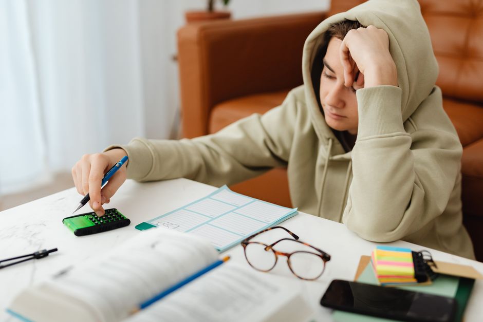 Teen wearing hoodie, using calculator at home, surrounded by study materials and glasses.