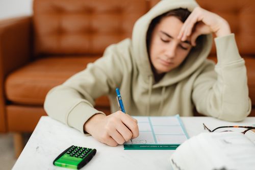 Free Teenage boy in a hoodie studying at home, visibly tired with a hand on his head. Stock Photo