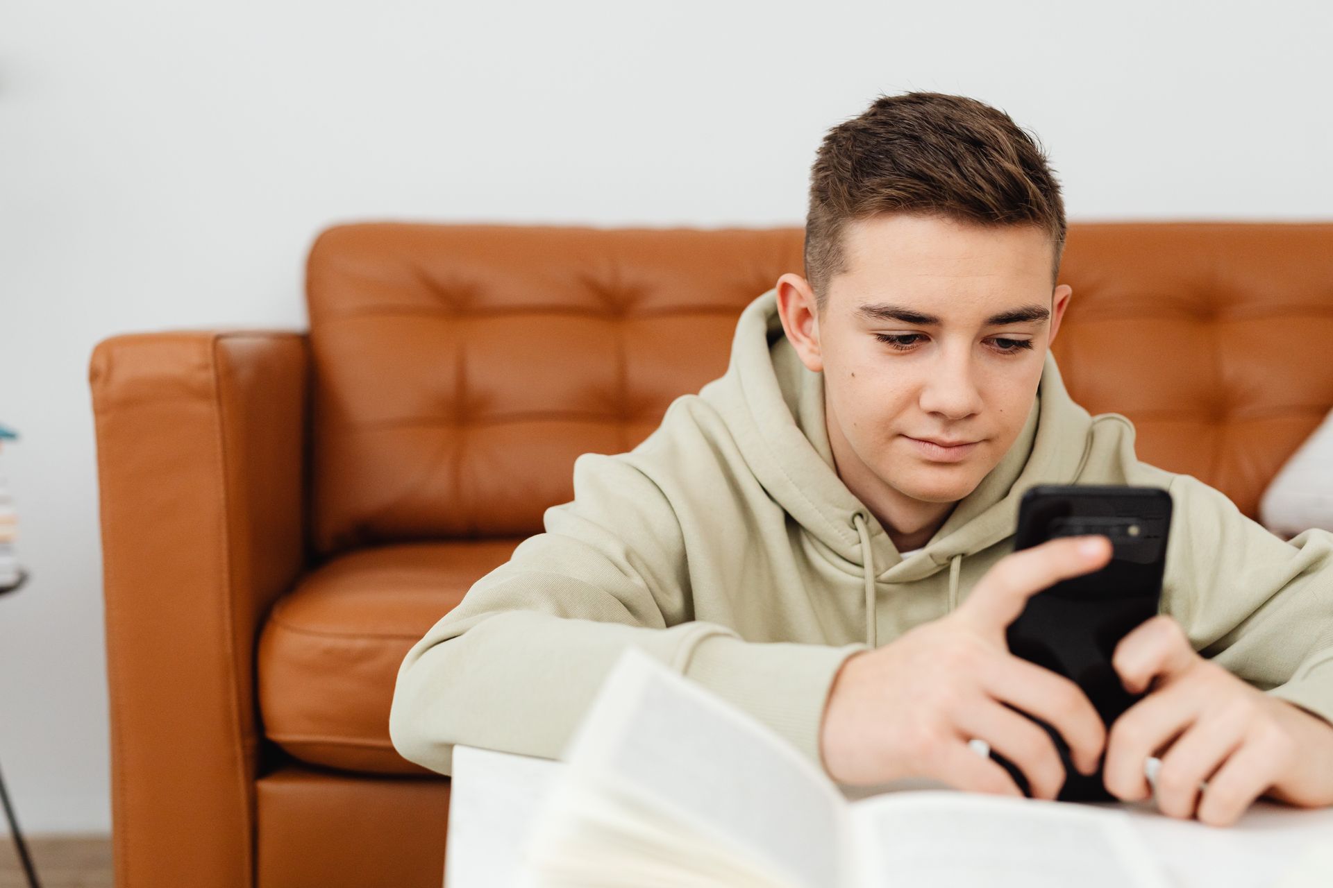 Teenager in a hoodie using a smartphone while sitting indoors with a book nearby.