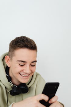 Happy teenager wearing hoodie and headphones, texting on a smartphone with a smile.