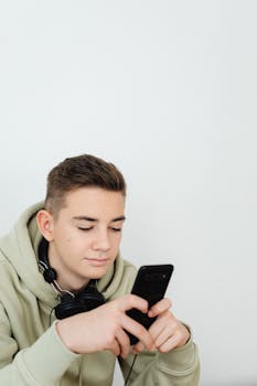 Teenage boy using a smartphone with headphones around his neck against a white background.