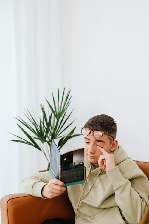 Free A young man in a hoodie reads a book indoors, adjusting his glasses for clarity. Stock Photo