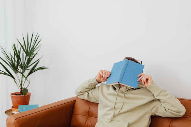 A Person Wearing A Hoodie Covering His Face With A Book