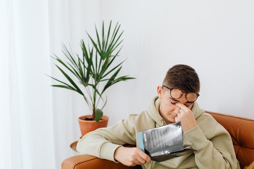 Free Adult man wearing hoodie rubs his eyes while reading a book indoors, next to a potted plant. Stock Photo
