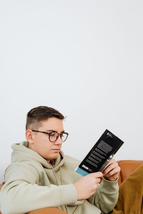 Free A young man in a hoodie reading a book indoors, showing focused concentration and relaxation. Stock Photo