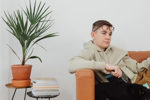 Free A young man sitting on a sofa with eyeglasses, next to a potted plant and stack of books. Stock Photo