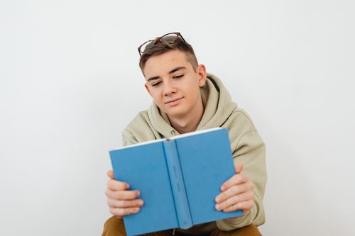 Free Teenager in a hoodie reading a blue book against a white background, conveying a relaxed study vibe. Stock Photo