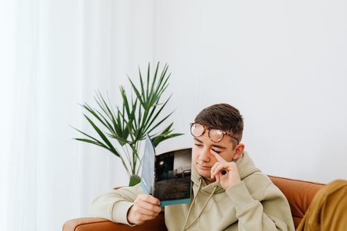 Free Young man wearing hoodie reading a magazine on a couch with a plant nearby. Stock Photo