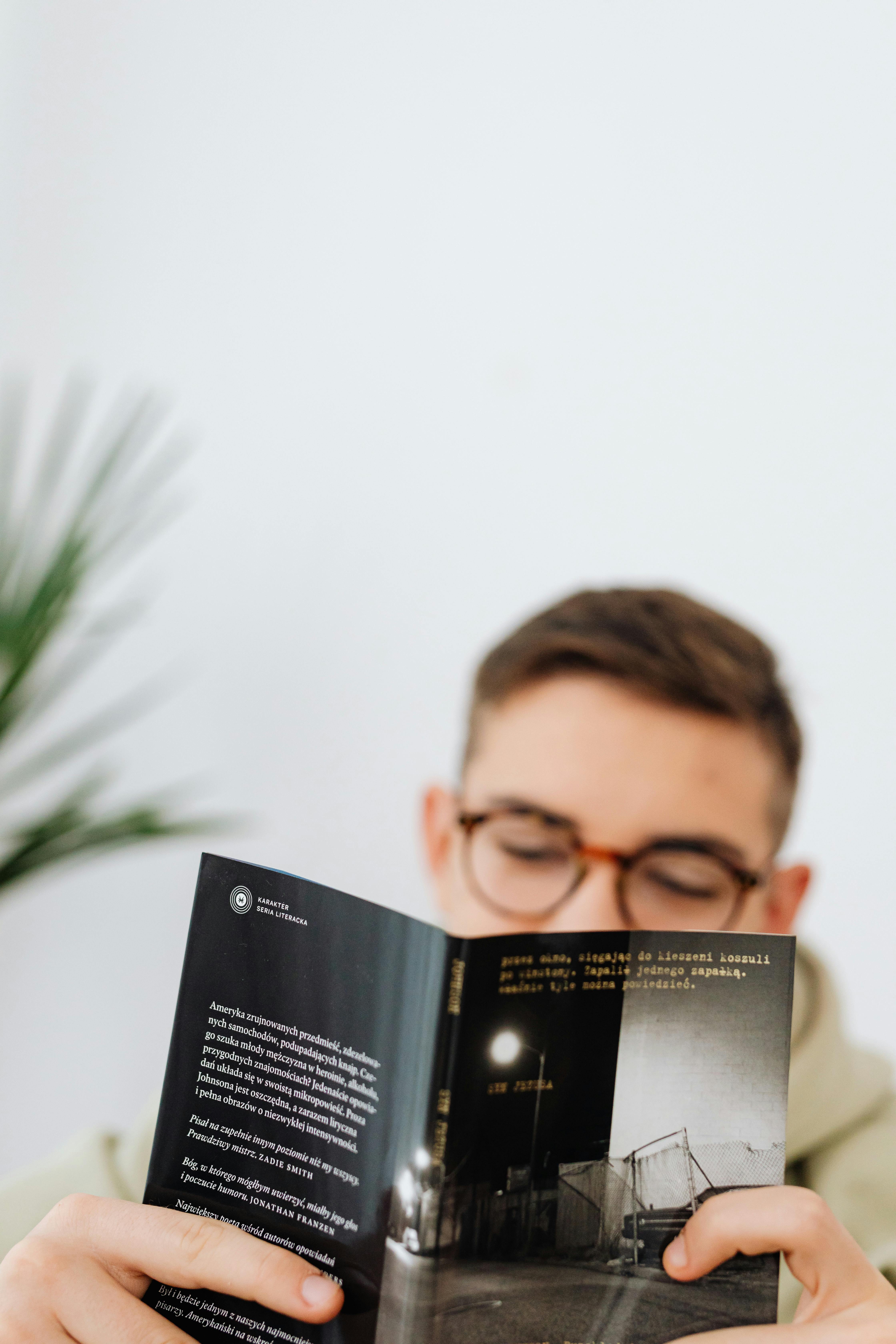 A young adult man wearing glasses reads a book indoors, focusing intently on the page.
