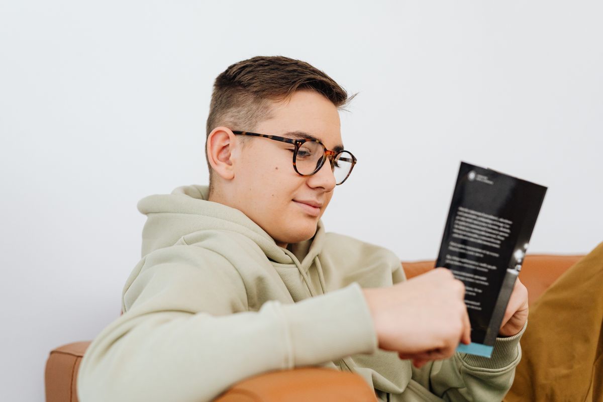 A young man wearing glasses reading a book while relaxing indoors