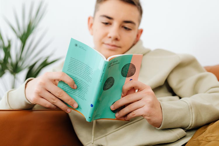 Teenage Boy Reading A Book And Sitting On A Couch 