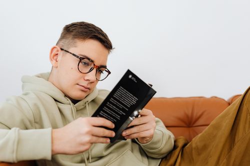 Free Young man wearing glasses and a hoodie, relaxing on a couch while reading a book. Stock Photo