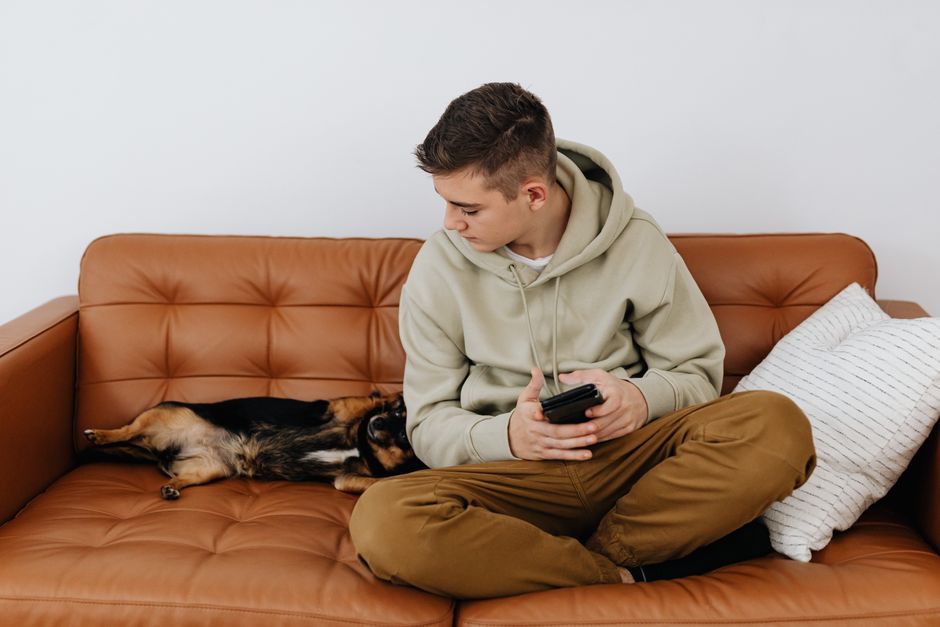 Teenager in casual attire with a smartphone, sitting on sofa with a dog.