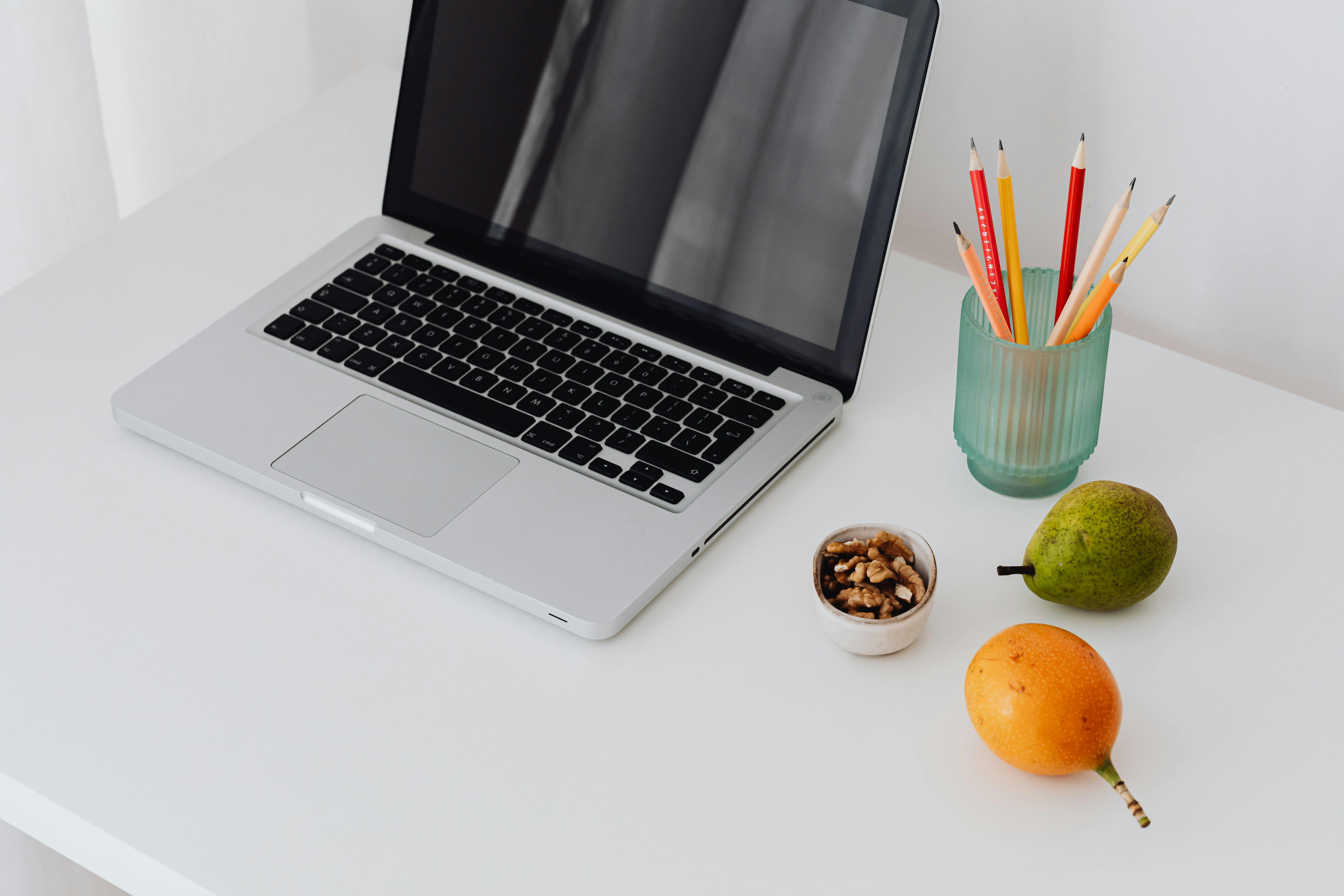 Simple office setup featuring a laptop, fruits, and stationery on a clean white desk.