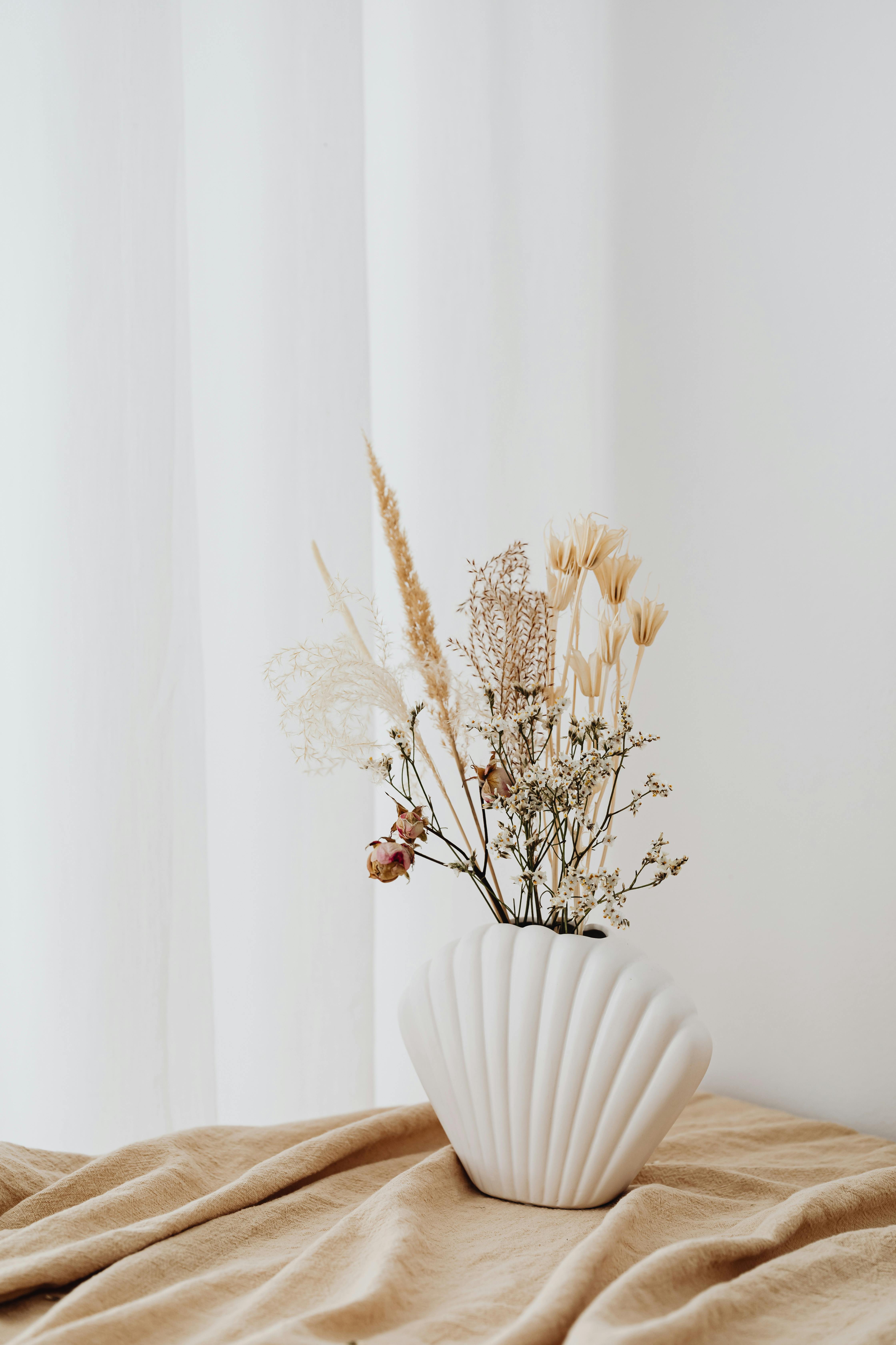 A close-up of an elegant white ceramic vase holding an arrangement of dried flowers on a tan fabric.