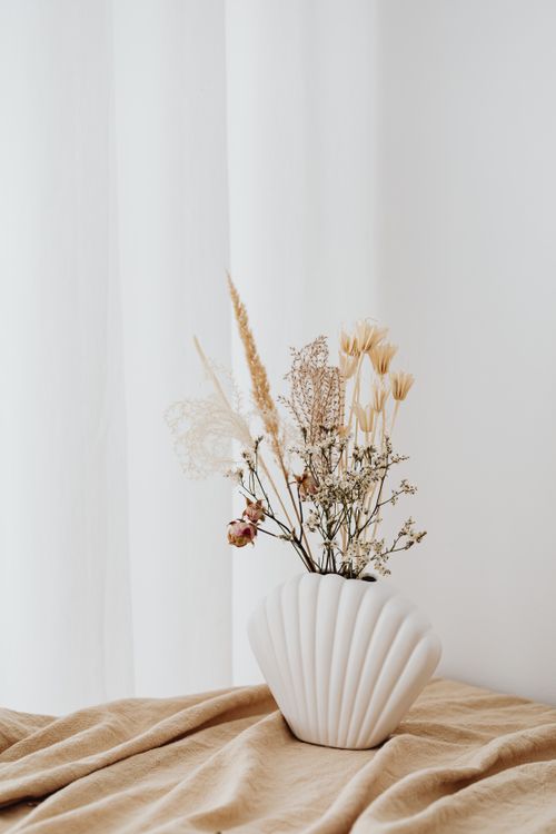 Free A close-up of an elegant white ceramic vase holding an arrangement of dried flowers on a tan fabric. Stock Photo