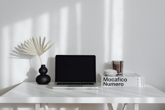 A simple and elegant workspace setup with a laptop, vase, and decor on a white table.
