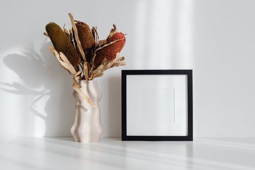Free A minimalist setup featuring dry flowers in a vase beside an empty picture frame. Stock Photo