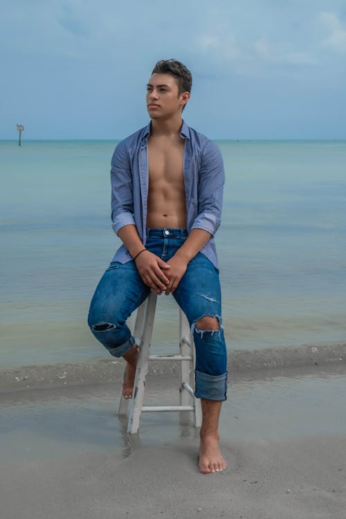 Full body of serious young man in jeans and denim shirt sitting on stool on sandy beach near ocean while looking away in daylight under gray cloudy sky