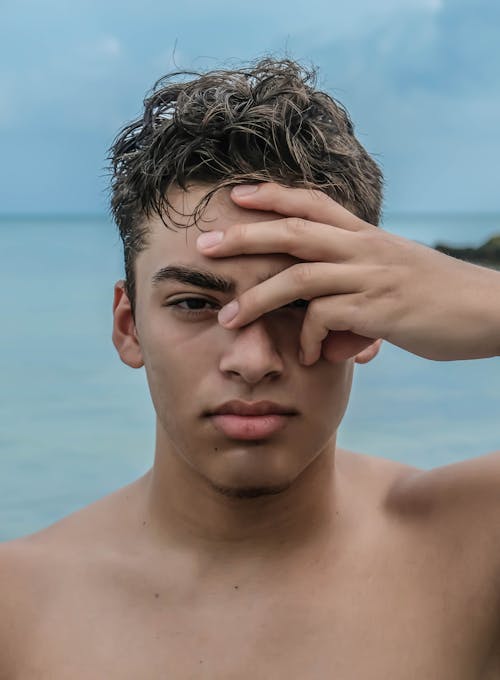 Serious young shirtless man covering eye with hand near ocean while looking at camera in daylight under gray cloudy sky
