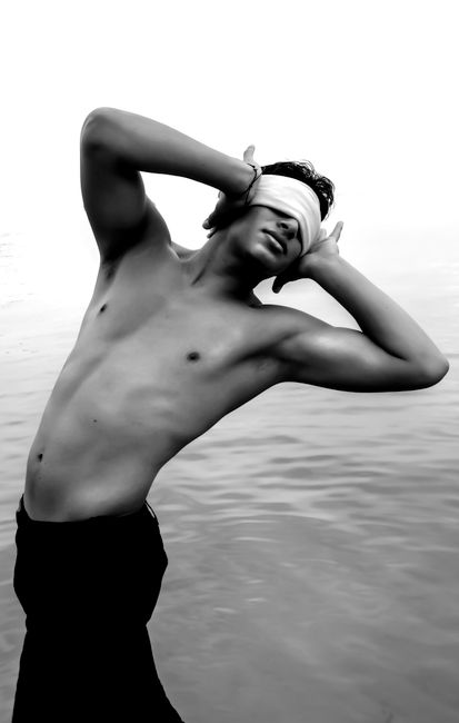 Black and white of pensive young topless male in jeans with blindfold on eyes covering ears with hands near rippling ocean in daytime