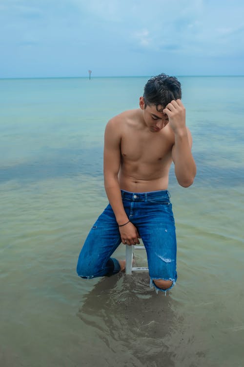 Full body of serious young man in jeans with naked torso sitting on stool in rippling ocean in daylight under gray cloudy sky