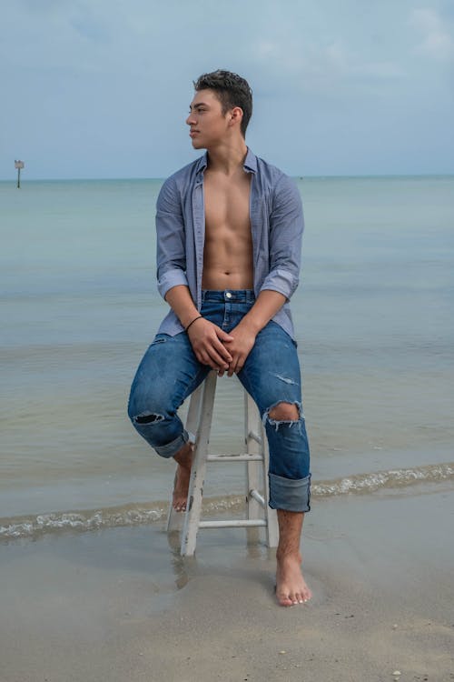 Full length of pensive young male in denim shirt and jeans sitting on stool on sandy beach near rippling ocean in daytime while looking away under gray sky