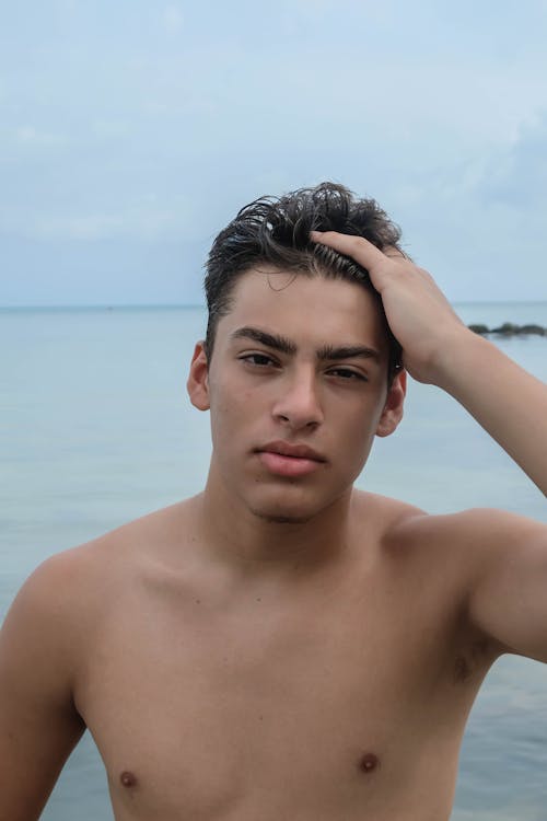 Free Pensive young shirtless male with hand on head looking at camera near rippling ocean in daytime under gray cloudy sky Stock Photo