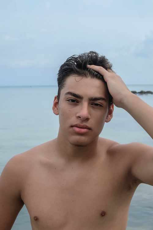 Free Pensive young shirtless male with hand on head looking at camera near rippling ocean in daytime under gray cloudy sky Stock Photo