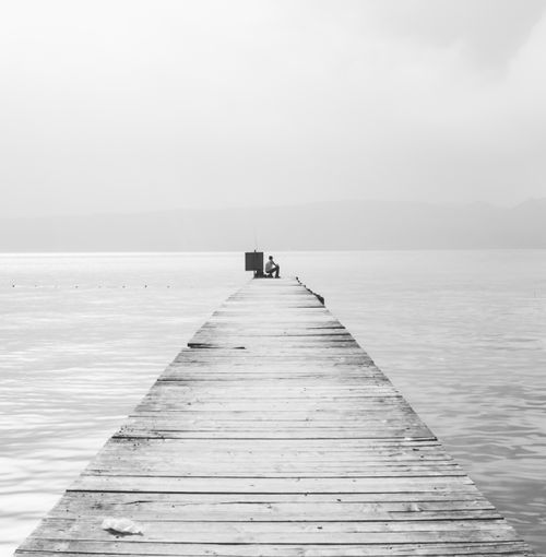 Free Serene monochromatic view of a lone figure fishing on a wooden boardwalk extending into the sea. Stock Photo