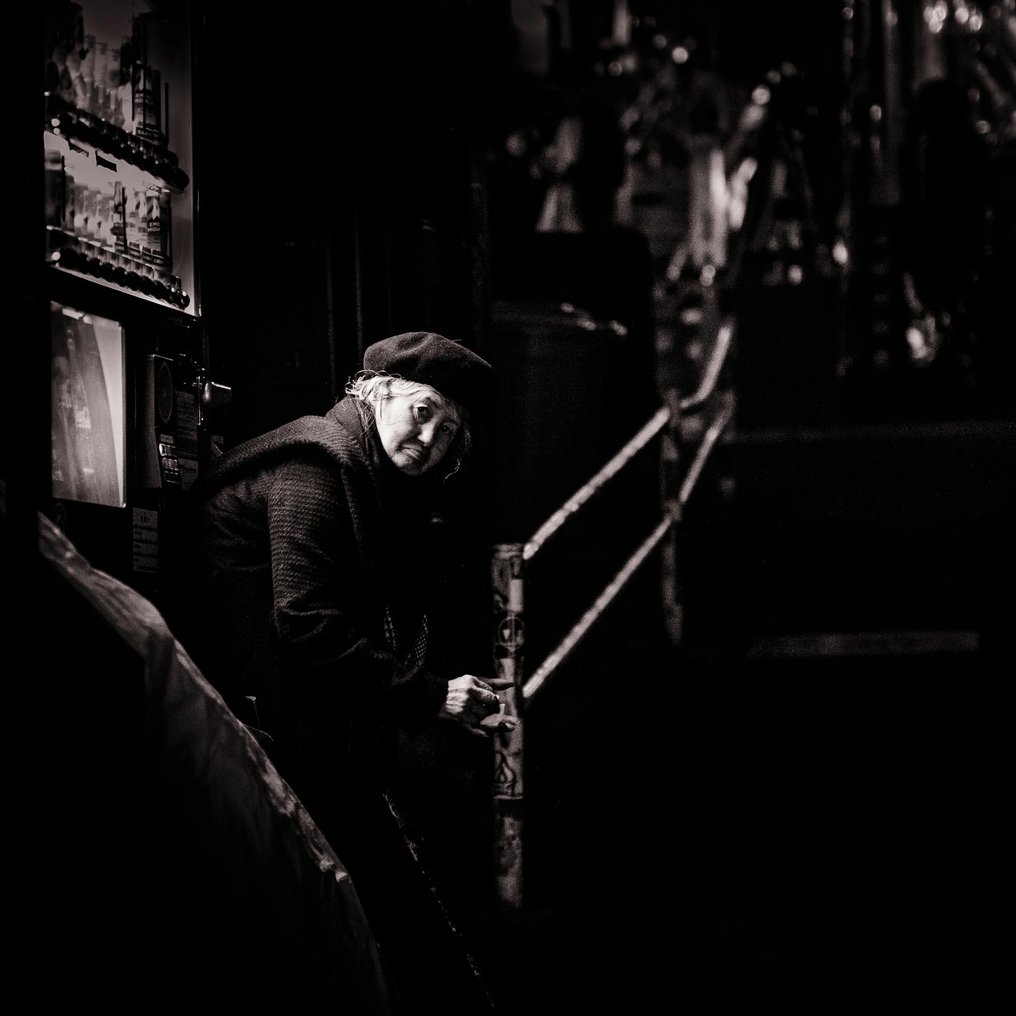 在一张黑白照片中，一位戴着贝雷帽的老妇人坐在光线昏暗的城市环境中