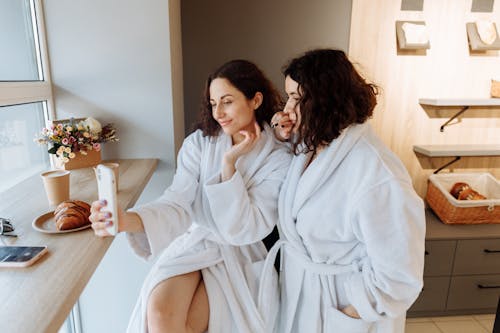Two women in bathrobes take a selfie with pastries and coffee in a cozy setting.