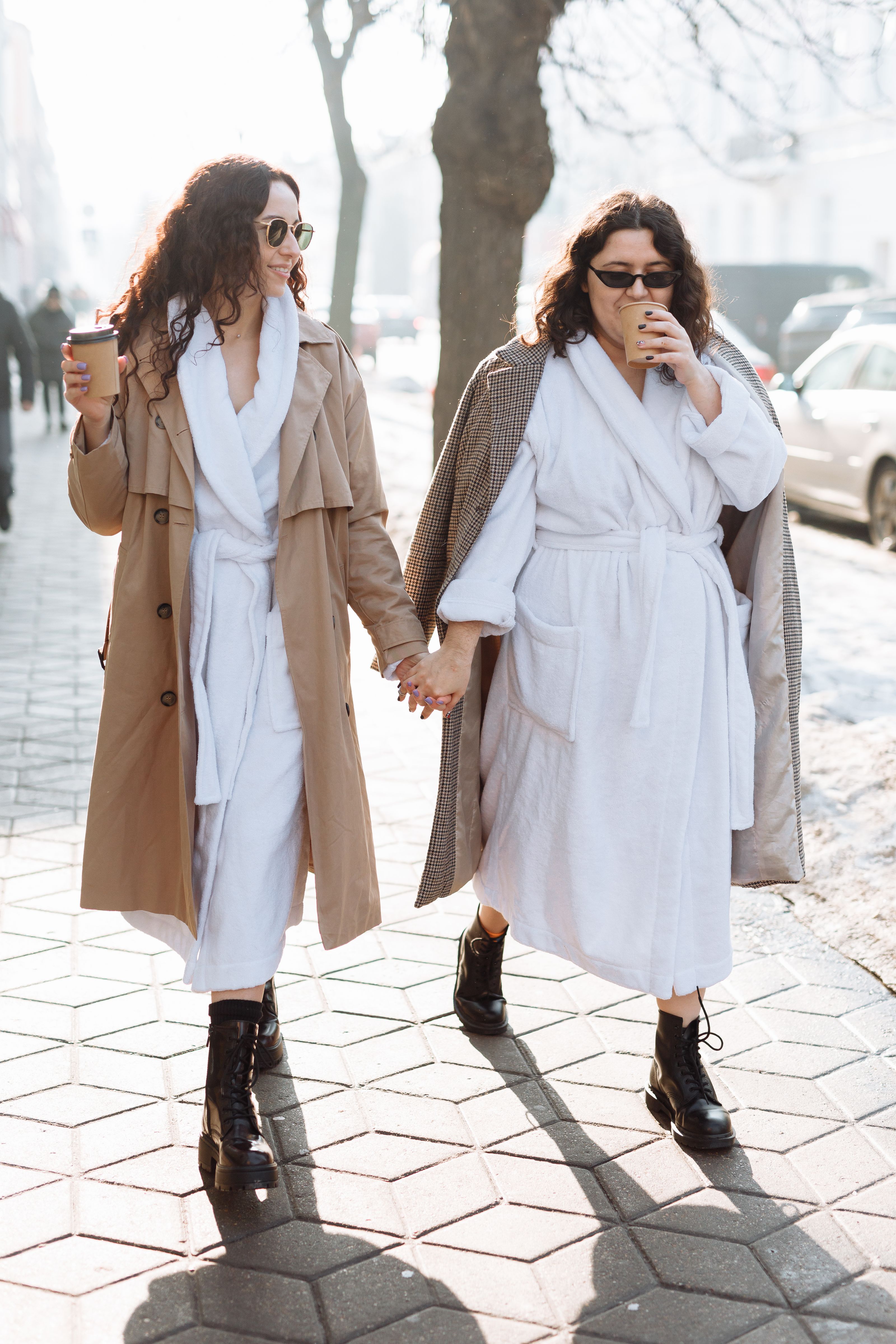 Duas mulheres em roupões segurando as mãos e caminhando ao ar livre, apreciando café.