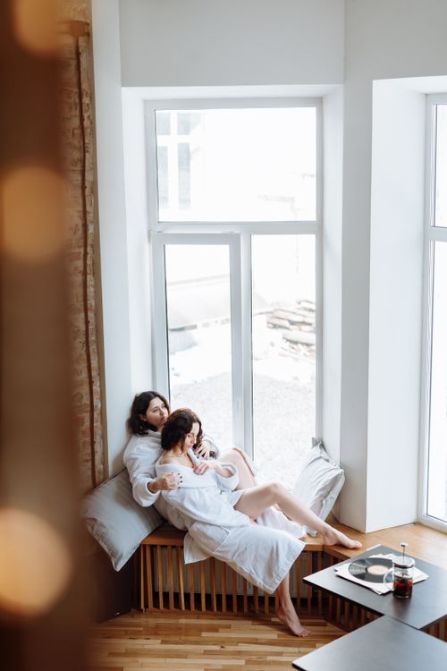 Free Two women in robes enjoying a peaceful moment by a window, embracing romance and togetherness. Stock Photo