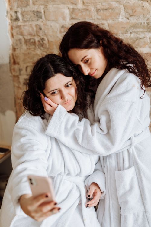 Free Two women embrace and take a selfie in cozy white bathrobes, reflecting a moment of affection. Stock Photo