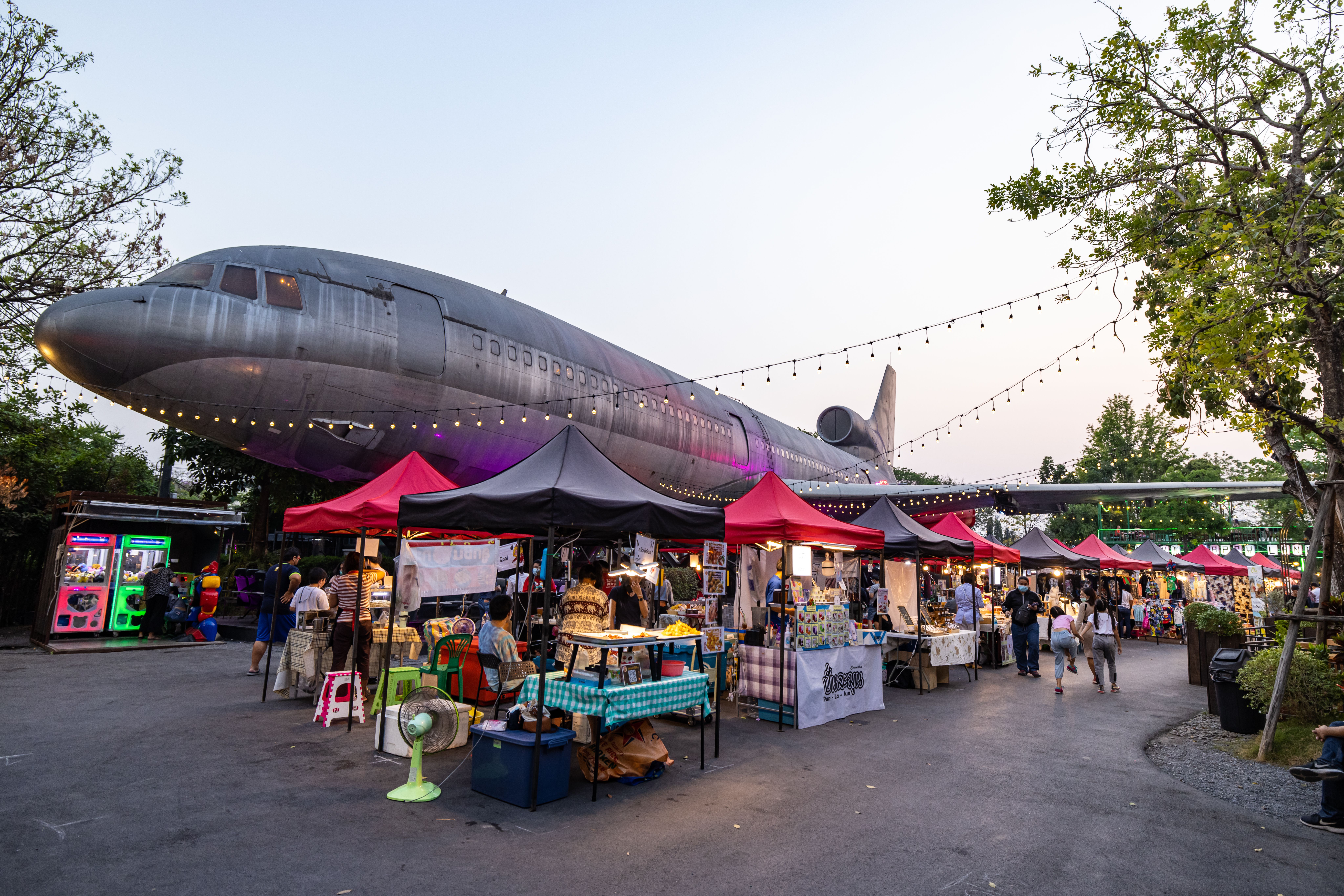 Bustling outdoor market scene with diverse stalls under an airplane, vibrant lights and evening atmo