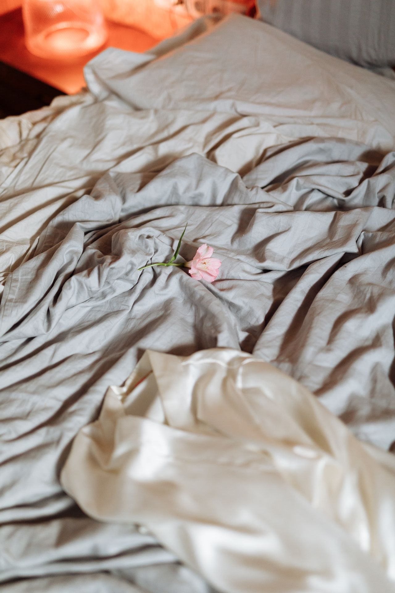 Crumpled grey-blue sheets with a pink flower resting on them beside a satin pillow