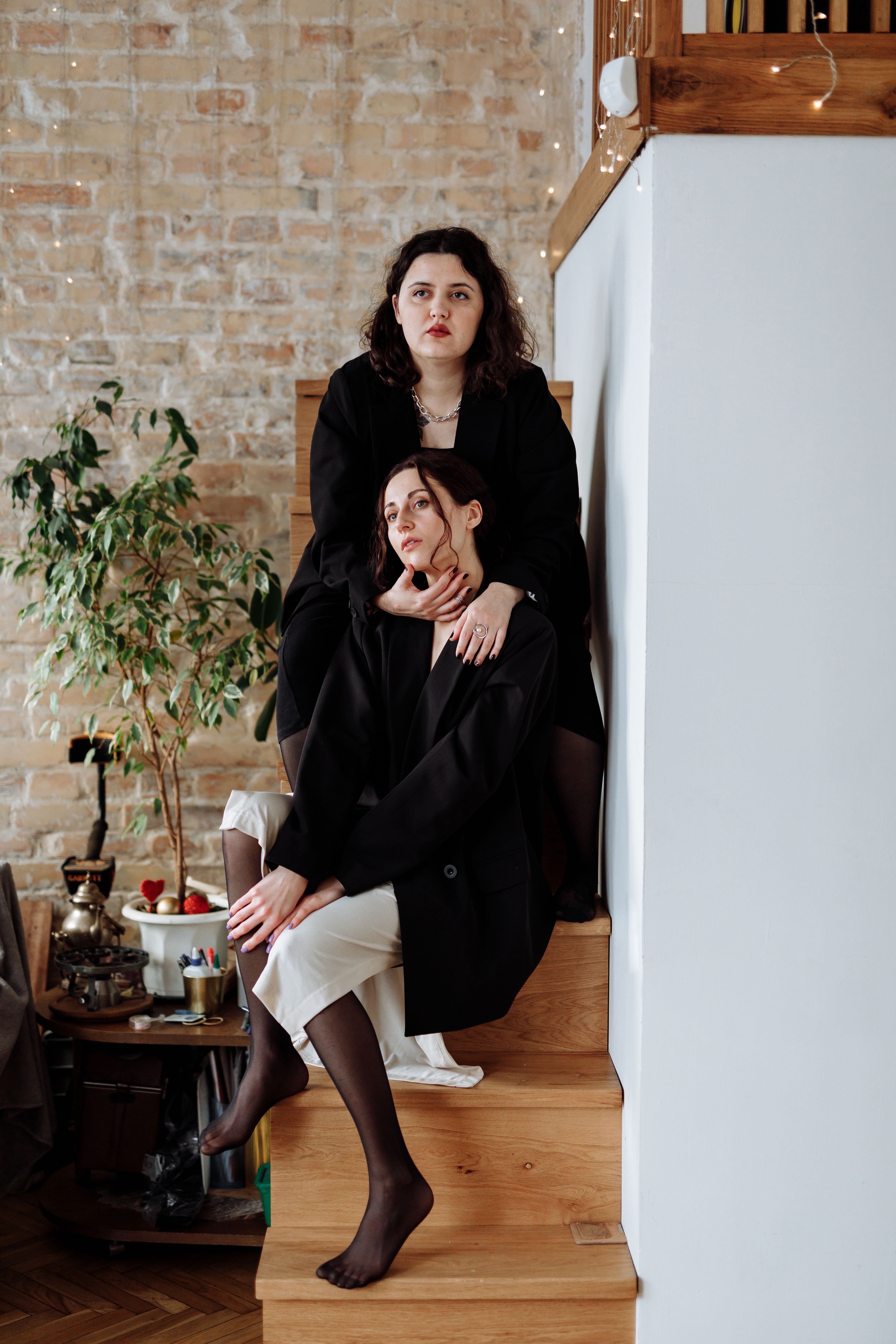 A couple in black coats sits together on wooden stairs indoors, embracing.