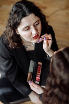 A woman in a black blazer applies lipstick to her partner indoors, highlighting intimacy and connection.