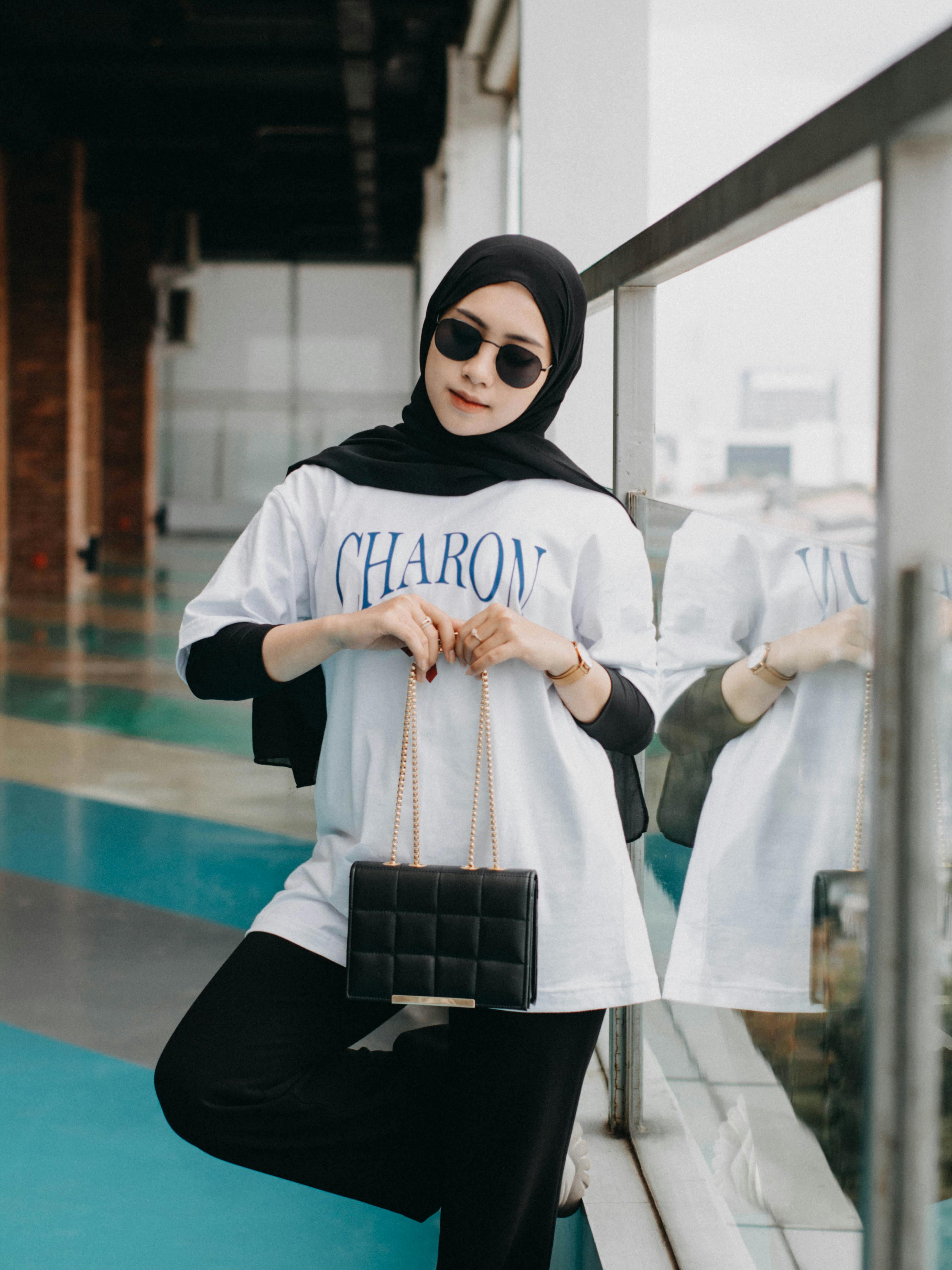 Fashionable woman in hijab with sunglasses and handbag posing indoors by glass railing.