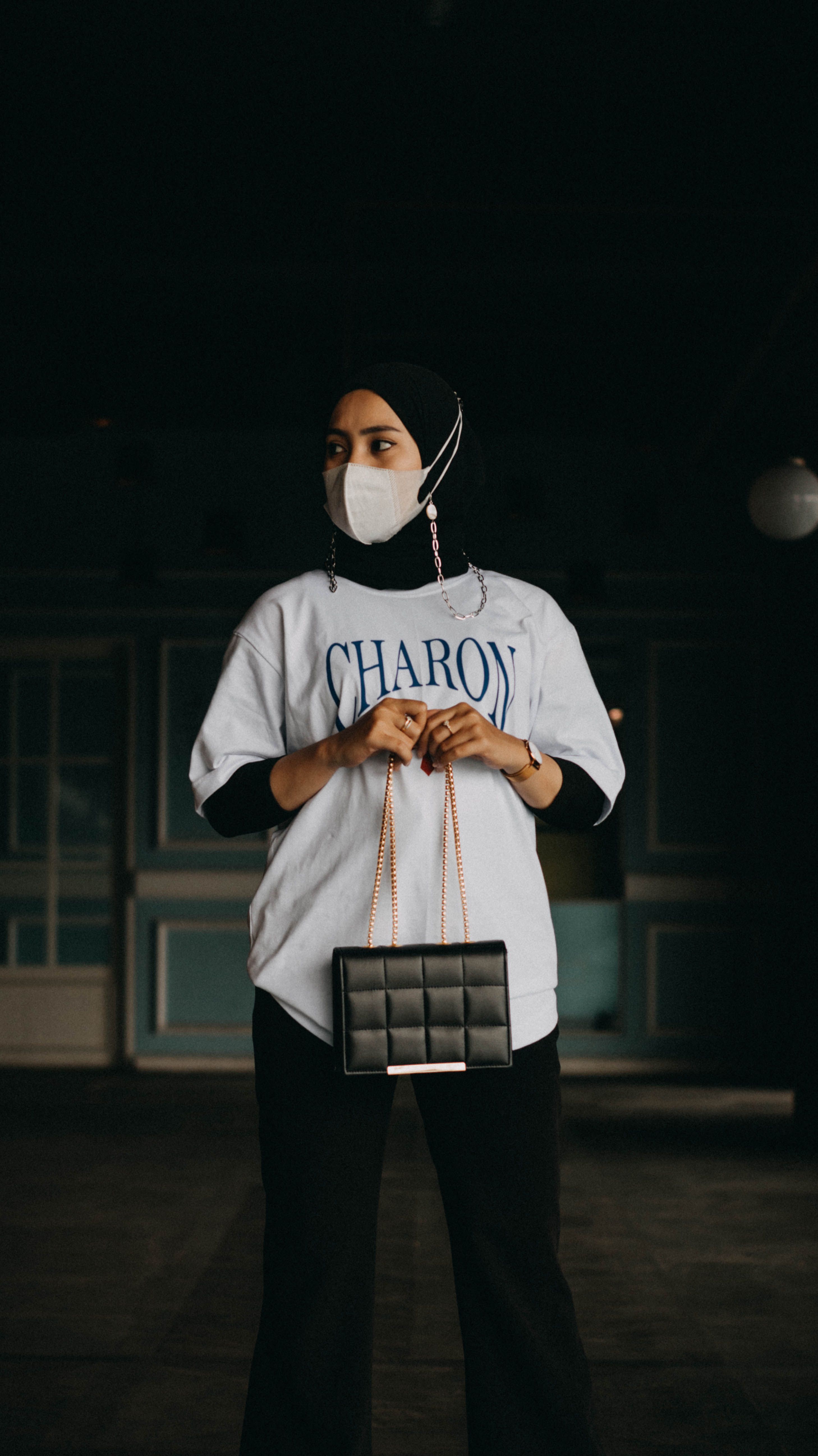 A woman standing indoors wearing a face mask, holding a trendy handbag, and wearing a hijab.