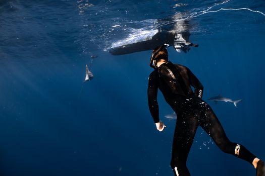 Back view anonymous male diver in wetsuit and snorkeling mask swimming in blue seawater near floating motorboat and fish