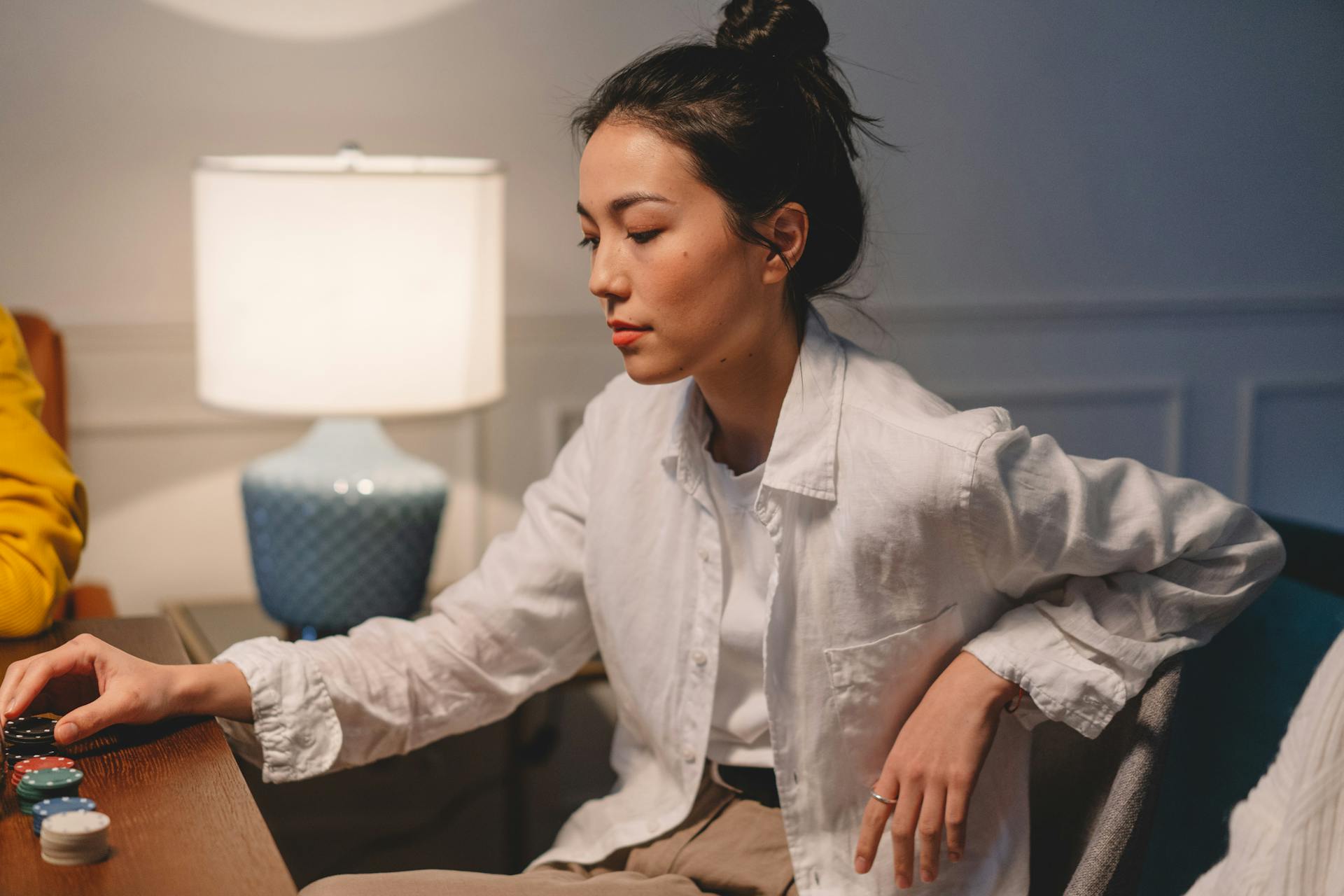 Woman in white shirt sitting by poker chips with lamp, focused on game.