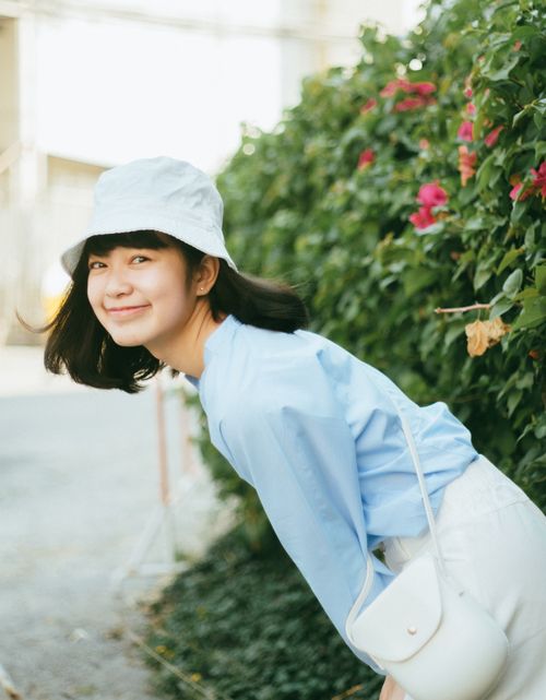 Free A cheerful woman with a hat poses in front of a lush green hedge outdoors in daylight. Stock Photo