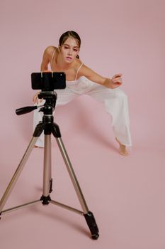 A fashion model poses creatively in a studio setting with a smartphone and tripod, against a pink background.