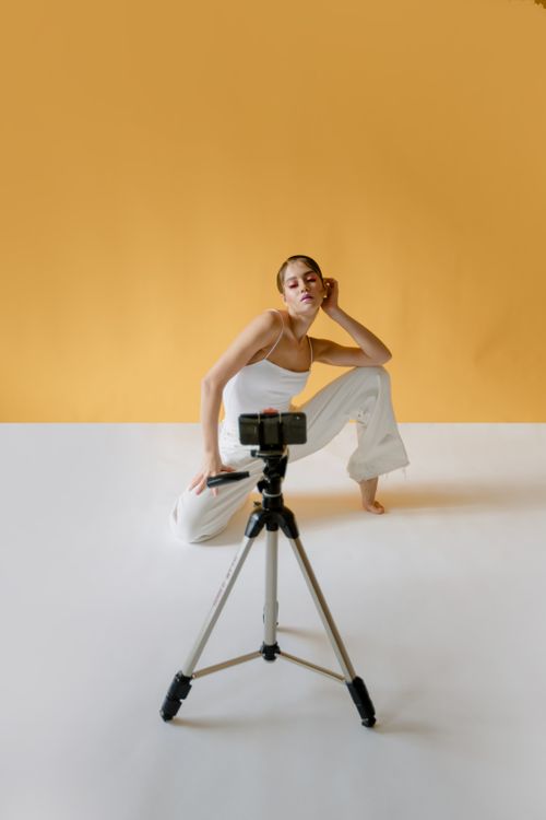 Free A stylish model poses in white clothes with a smartphone on a tripod against a yellow background. Stock Photo