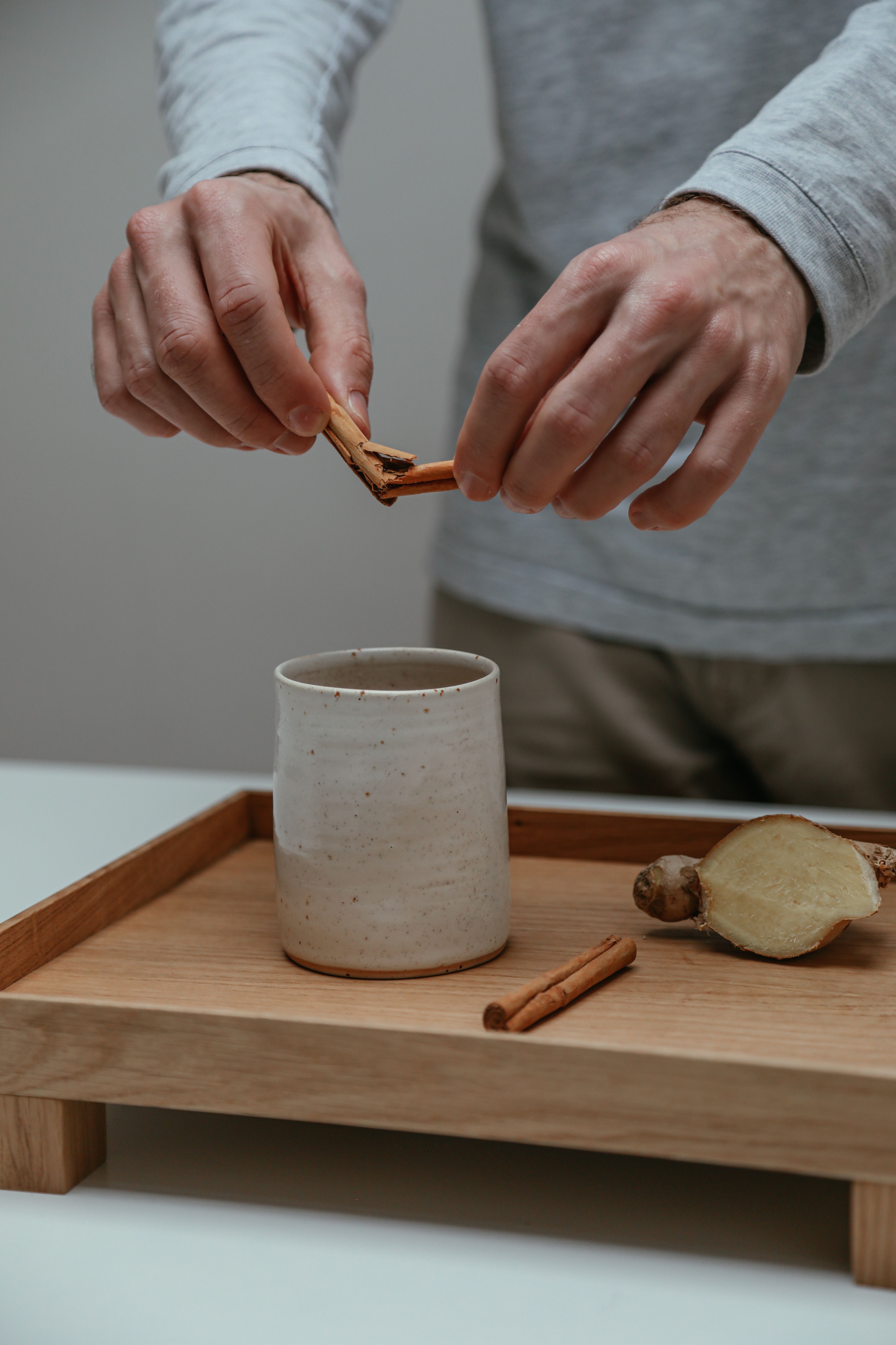 Hands placing cinnamon sticks into a cup of tea on a wooden tray with ginger.