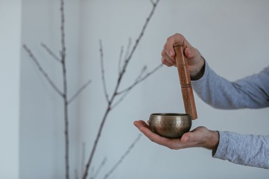 Hands holding a brass singing bowl with a wooden mallet against a minimalist indoor background.