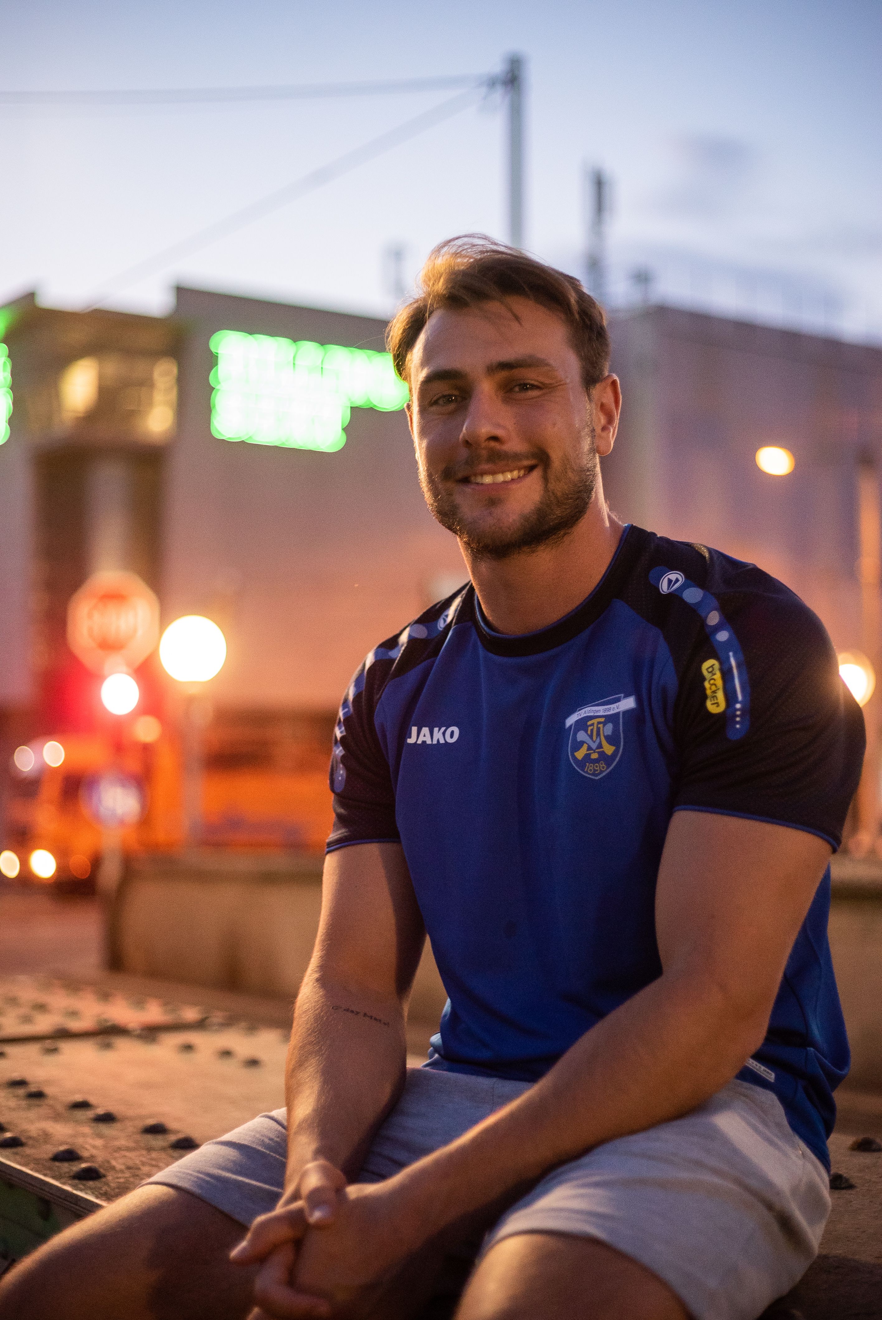 A man sitting and smiling outdoors during the evening in Stuttgart, Germany, wearing a blue sports shirt.