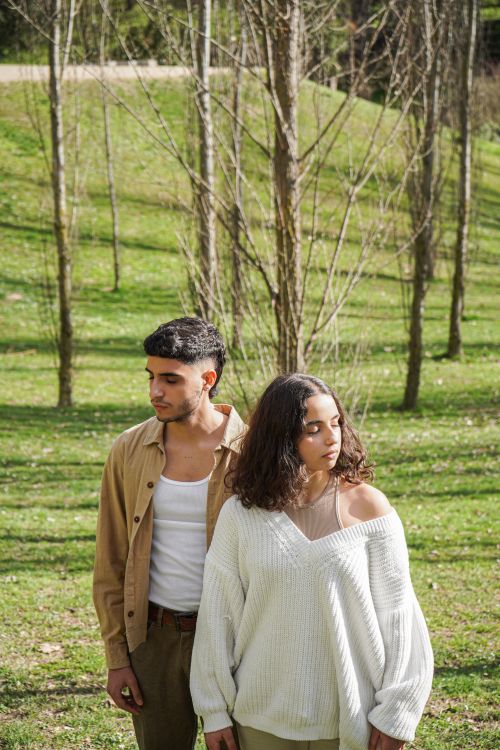 Free A young couple stands in a serene park, surrounded by trees, basking in the sunlight. Stock Photo